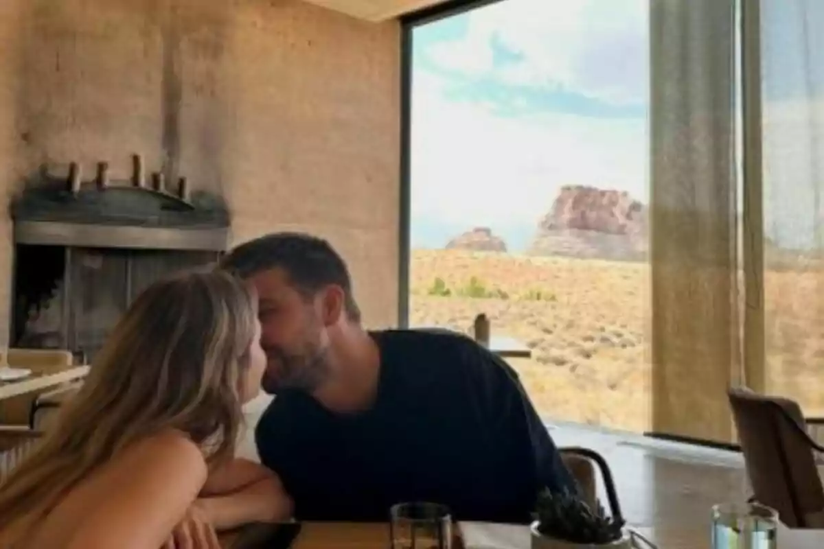 Parella asseguda en una taula de restaurant al costat d’una finestra gran amb vistes al desert i formacions rocoses mentre s’acosten per fer-se un petó. Parella asseguda en una taula de restaurant al costat d’una finestra gran amb vistes al desert i formacions rocoses mentre s’acosten per fer-se un petó.