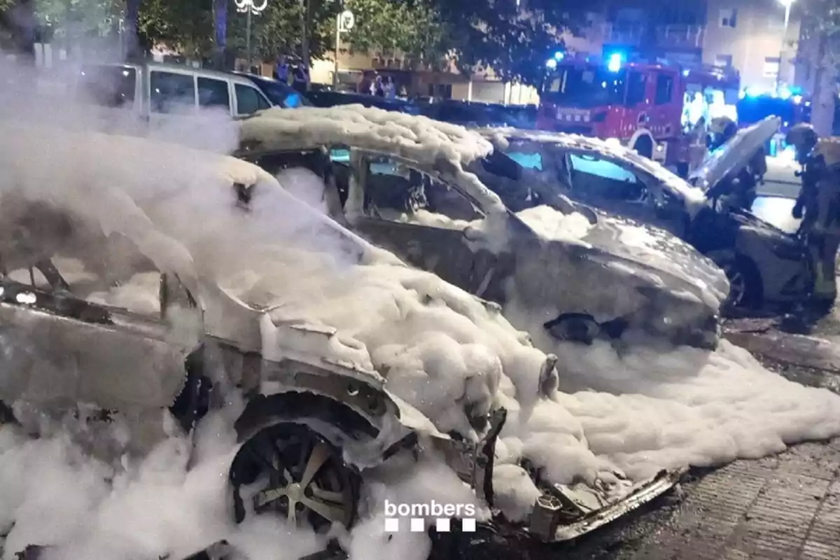 Diversos cotxes coberts d’escuma després d’un incendi a la via pública amb bombers i camions d’emergència al fons Diversos cotxes coberts d’escuma després d’un incendi a la via pública amb bombers i camions d’emergència al fons