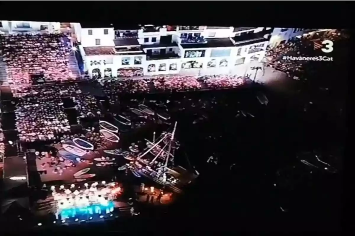 Vista nocturna d’un esdeveniment multitudinari a l’aire lliure vora el mar amb un escenari il·luminat, diverses barques i un vaixell decoratiu, envoltat de cases blanques i una gran quantitat de públic Vista nocturna d’un esdeveniment multitudinari a l’aire lliure vora el mar amb un escenari il·luminat, diverses barques i un vaixell decoratiu, envoltat de cases blanques i una gran quantitat de públic