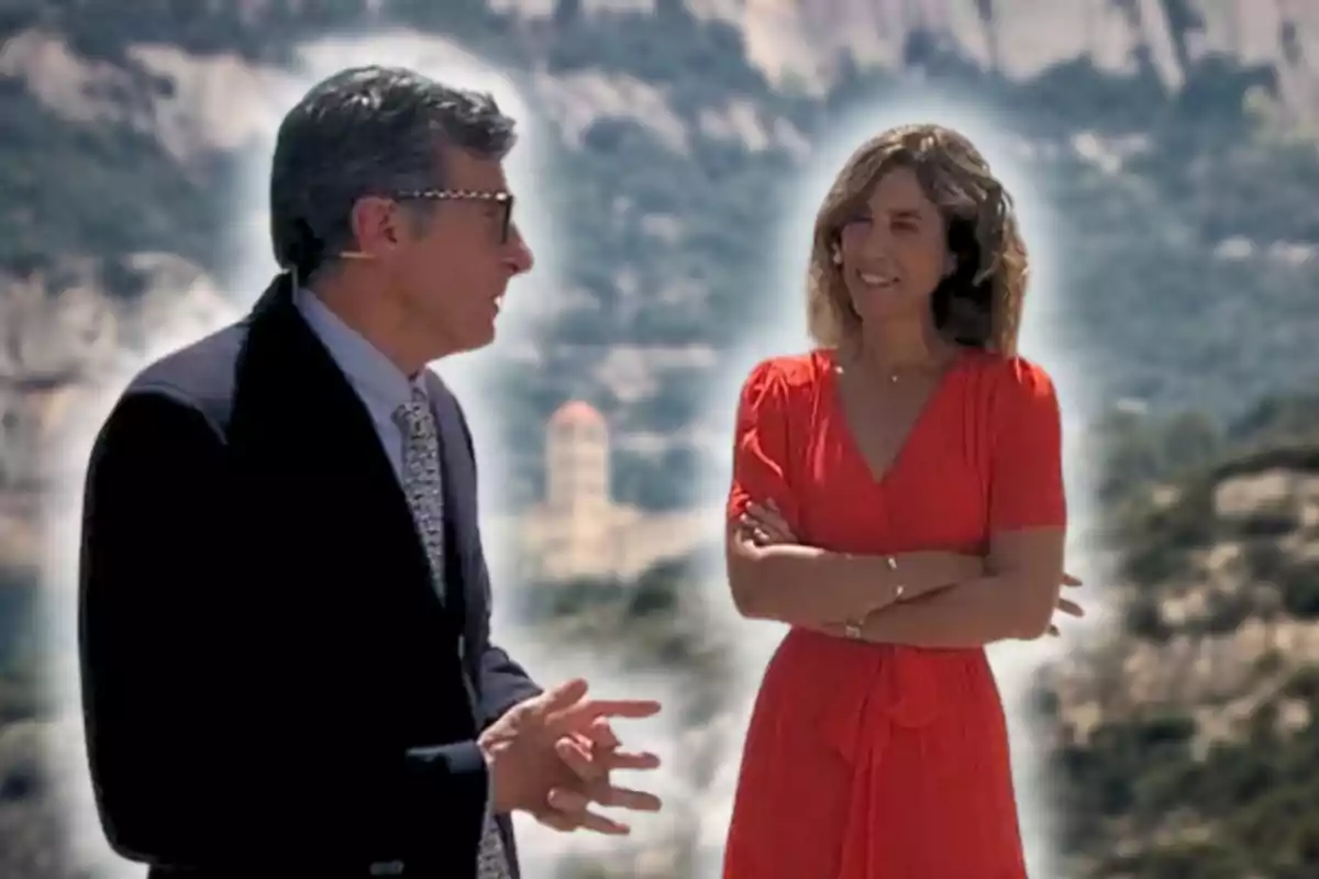Un hombre con gafas y traje habla con una mujer de vestido rojo que sonríe con los brazos cruzados, ambos están al aire libre con montañas de fondo.