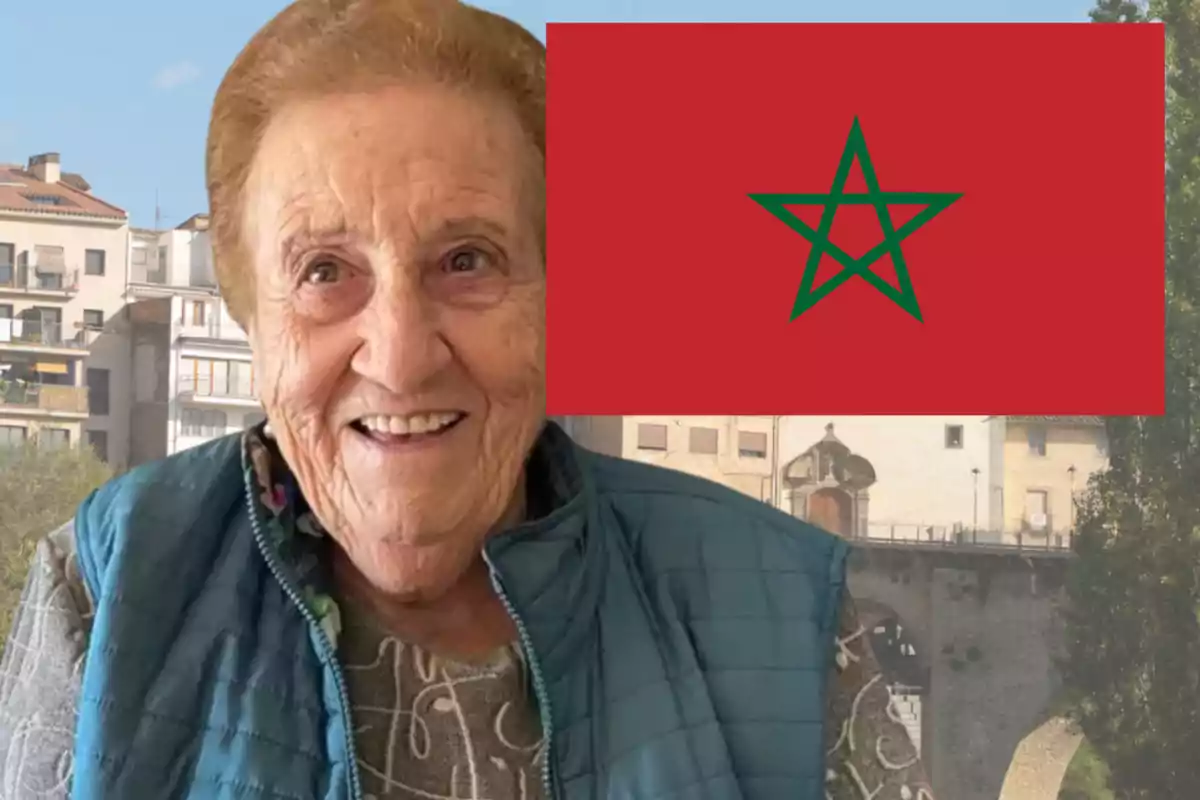 Mujer mayor sonriente con chaleco azul frente a una bandera de Marruecos y edificios al fondo
