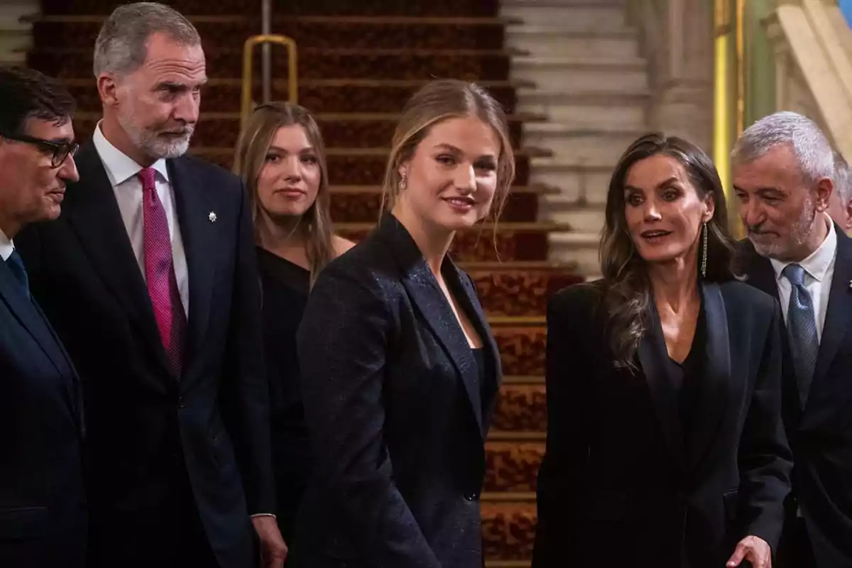 Grup de persones elegantment vestides conversant i somrient a l’interior d’un edifici amb una escala enmoquetada al fons