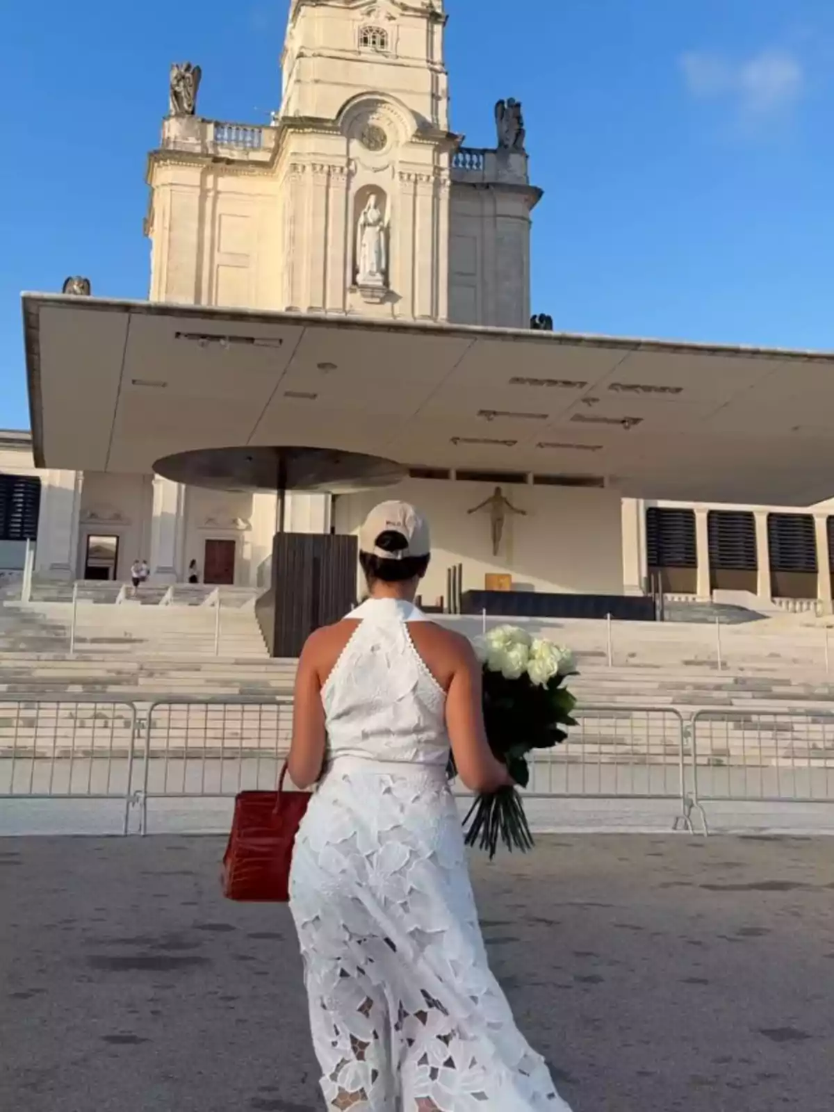 Georgina vestida de blanco con un ramo de flores y bolso rojo caminando hacia una iglesia grande bajo un cielo azul Georgina vestida de blanco con un ramo de flores y bolso rojo caminando hacia una iglesia grande bajo un cielo azul