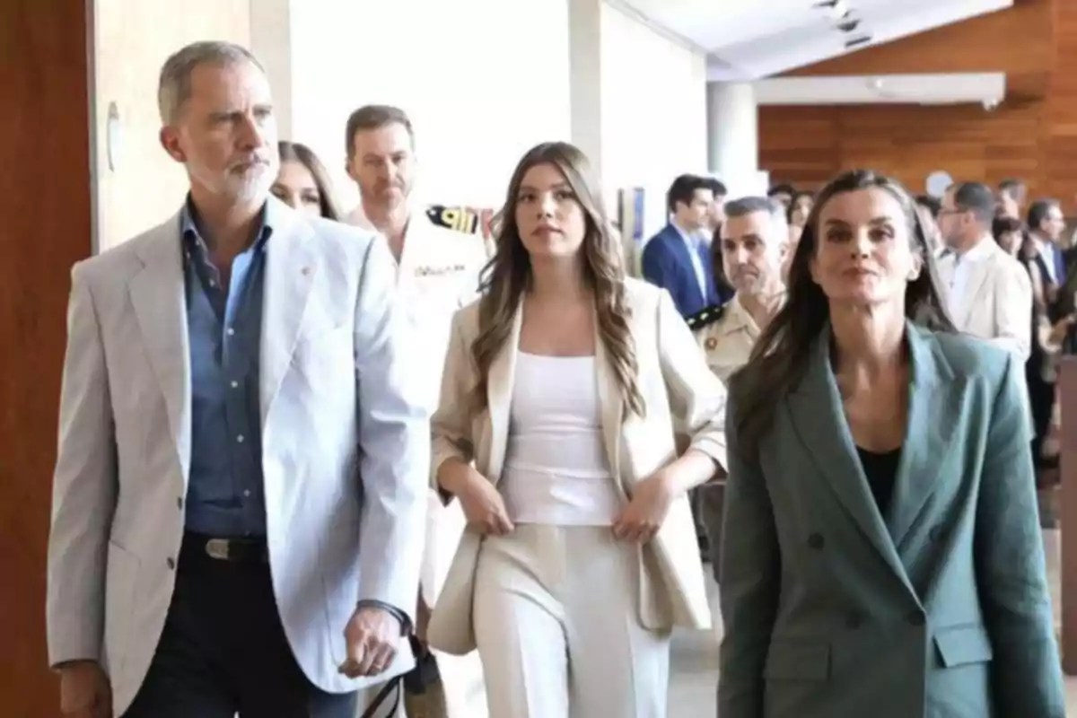 Three formally dressed people walk down a hallway accompanied by others in the background. Three formally dressed people walk down a hallway accompanied by others in the background.