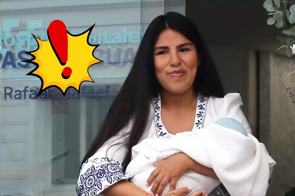 Isa Pantoja de cabello largo y oscuro sonríe mientras sostiene a un bebé envuelto en una manta blanca, con un gráfico de exclamación amarillo y rojo sobre el lado izquierdo de la imagen.