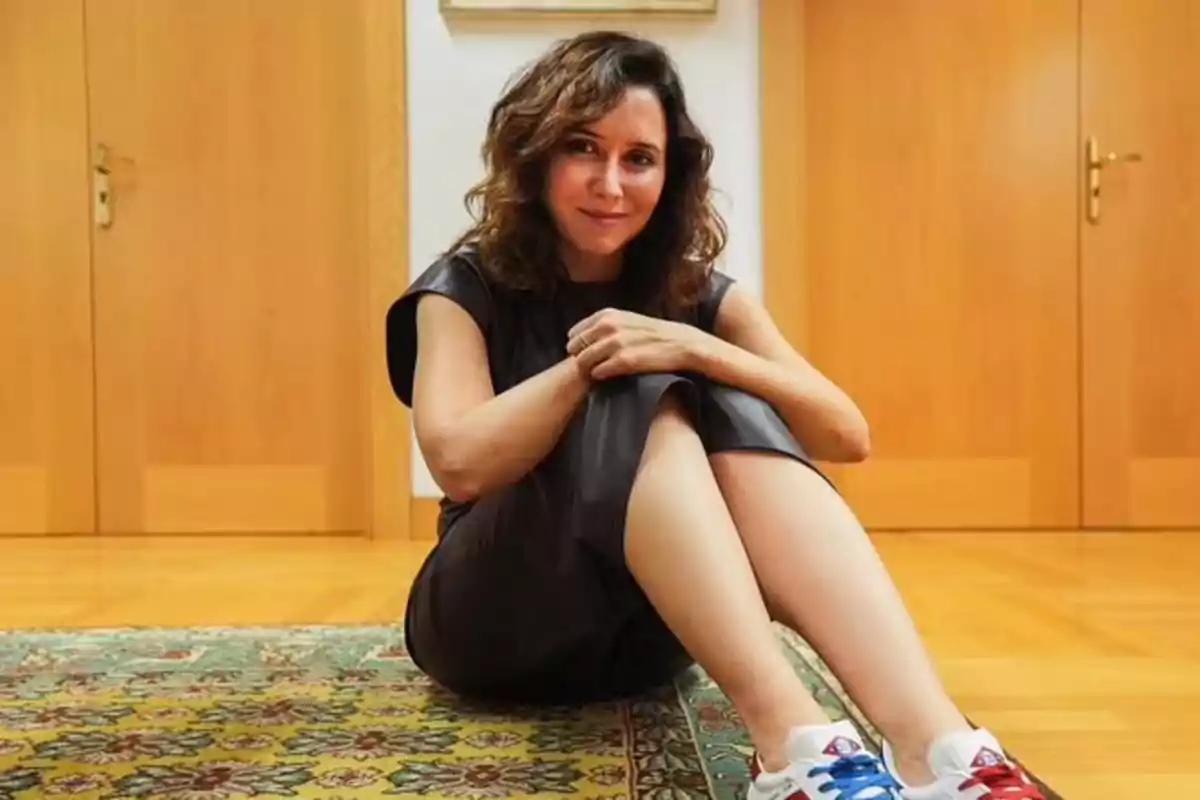 Isabel Díaz Ayuso sentada en el suelo sobre una alfombra colorida con las piernas cruzadas, lleva un vestido oscuro y zapatillas deportivas, al fondo hay puertas de madera.