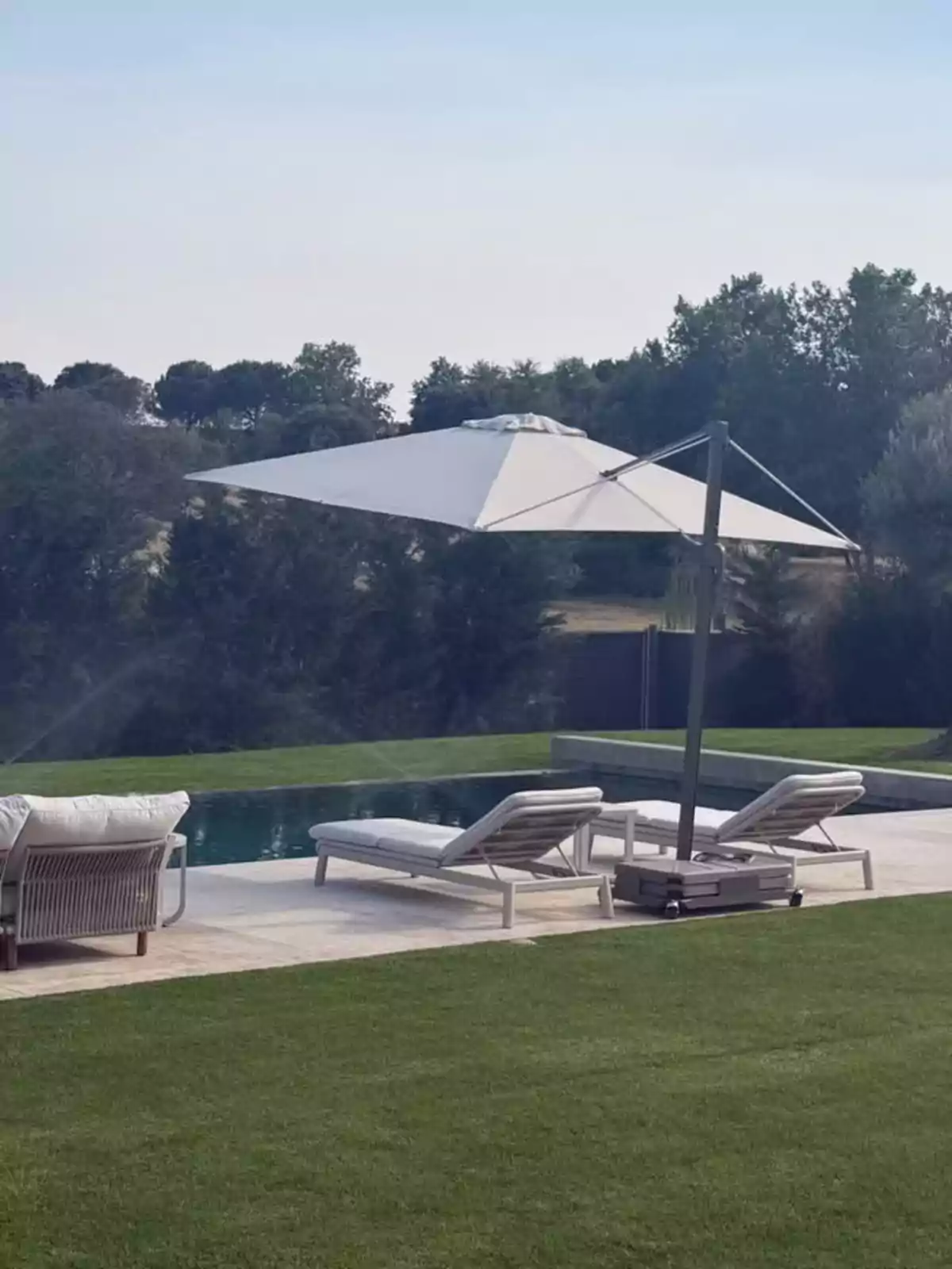 Dos tumbonas blancas junto a una piscina con una sombrilla grande en un jardín rodeado de árboles Dos tumbonas blancas junto a una piscina con una sombrilla grande en un jardín rodeado de árboles