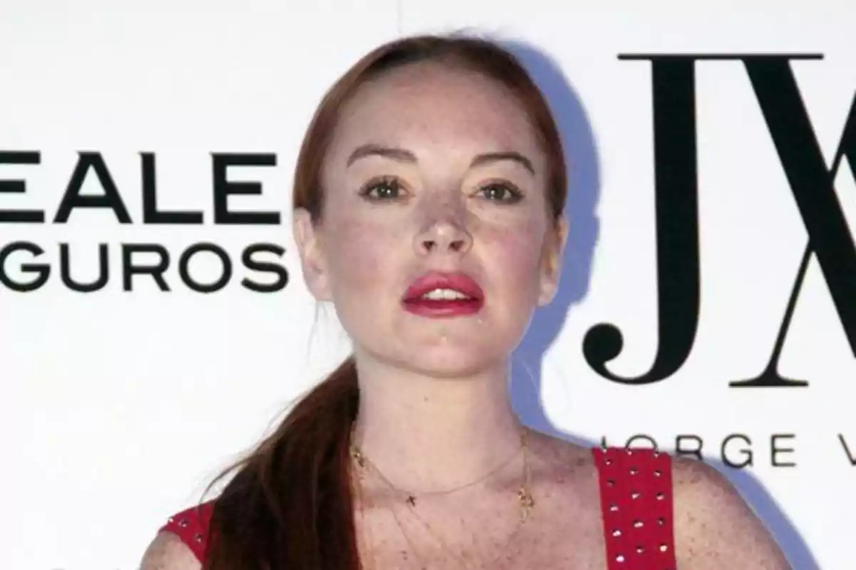 Lindsay Lohan pelirroja con vestido rojo y labios pintados posando frente a un fondo con logotipos de marcas Lindsay Lohan pelirroja con vestido rojo y labios pintados posando frente a un fondo con logotipos de marcas