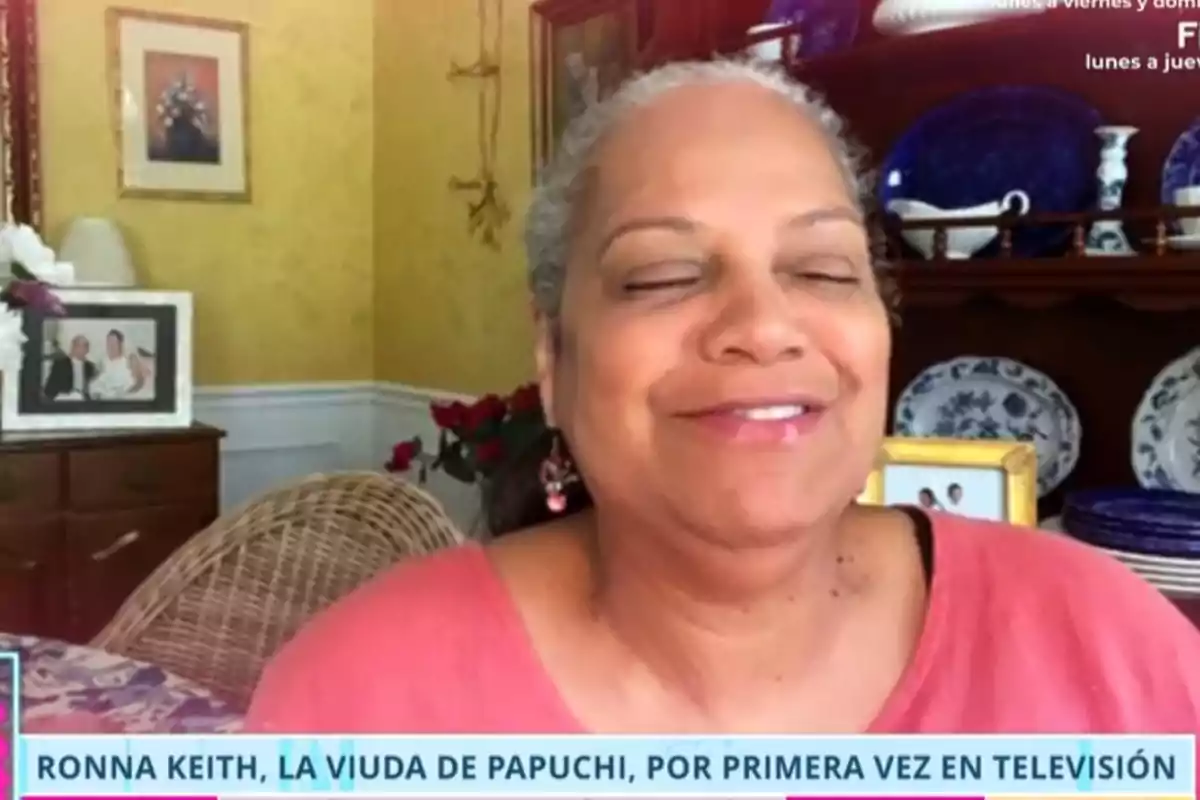 Ronna Keith sonriente con los ojos cerrados en una videollamada desde una sala decorada con platos y flores, con un cuadro y una foto familiar al fondo Ronna Keith sonriente con los ojos cerrados en una videollamada desde una sala decorada con platos y flores, con un cuadro y una foto familiar al fondo