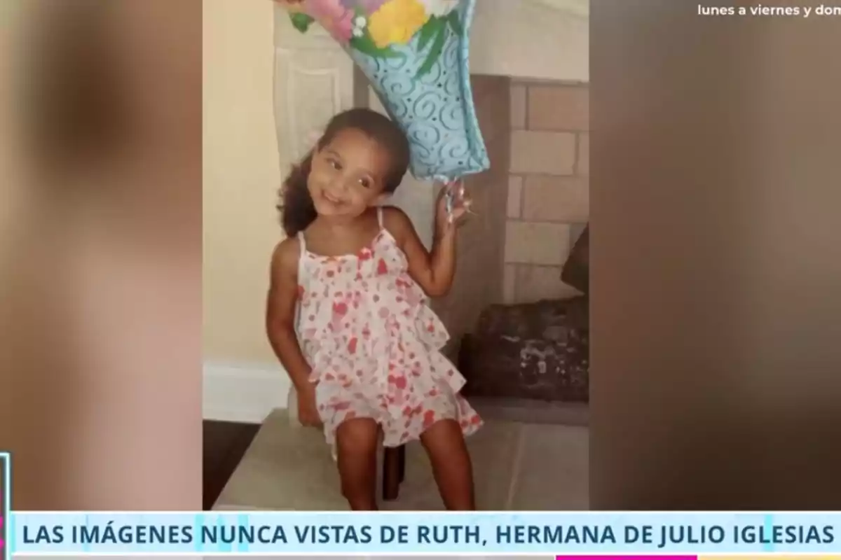 Ruth pequeña con vestido rosa y flores sentada en una silla sosteniendo un globo azul en forma de estrella frente a una chimenea Ruth pequeña con vestido rosa y flores sentada en una silla sosteniendo un globo azul en forma de estrella frente a una chimenea