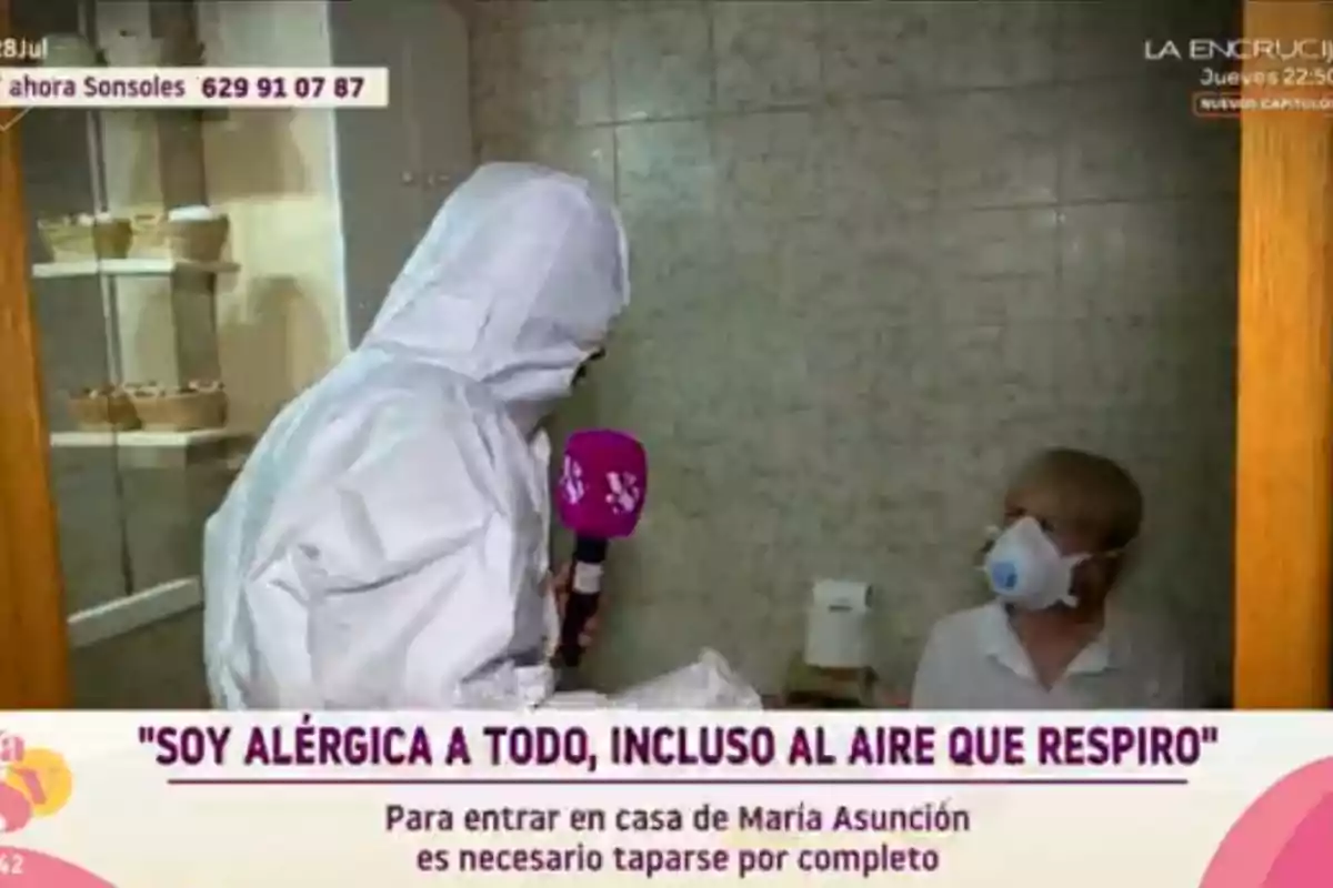 Una reportera con traje protector entrevista a una mujer que lleva mascarilla en una casa, mientras en la pantalla aparece un texto sobre alergias extremas. Una reportera con traje protector entrevista a una mujer que lleva mascarilla en una casa, mientras en la pantalla aparece un texto sobre alergias extremas.