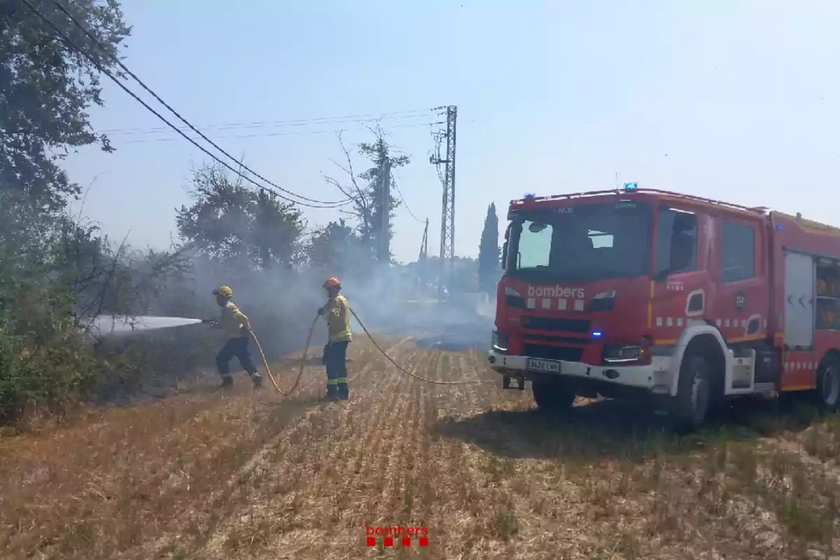Dos bombers apaguen un incendi en un camp sec amb una mànega d’aigua mentre un camió de bombers vermell està estacionat a prop.