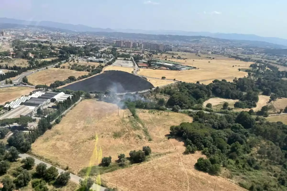 Vista aérea de un campo con vegetación seca y un área quemada con humo, rodeado de caminos, árboles y edificios al fondo