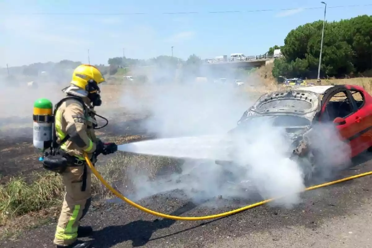 Bombarder apaga l’incendi d’un cotxe vermell en una carretera mentre surt fum del vehicle.