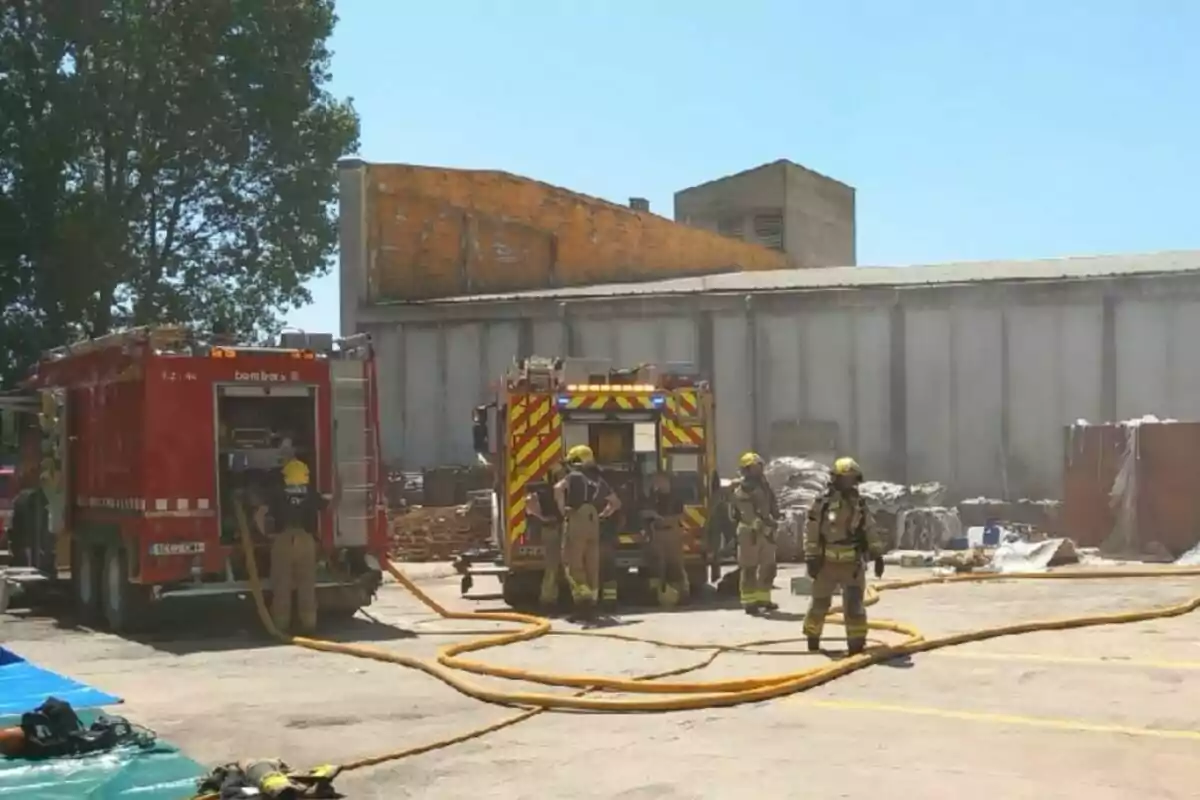 Bombers amb equip de protecció treballen al costat de dos camions vermells davant d’una nau industrial, amb mànegues esteses a terra i fum sortint de l’edifici.