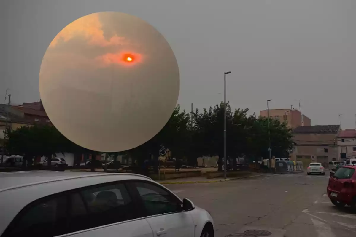 Calle de un pueblo con cielo nublado y el sol rojizo visible a través del humo, con varios coches estacionados y árboles alrededor de una plaza