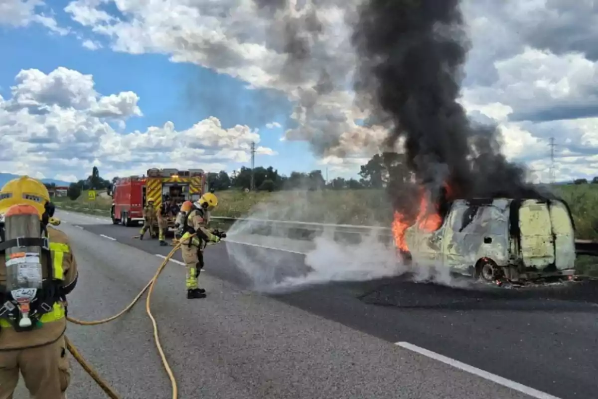 Bombers apaguen l’incendi d’una furgoneta en una carretera mentre una columna de fum negre s’eleva cap al cel.