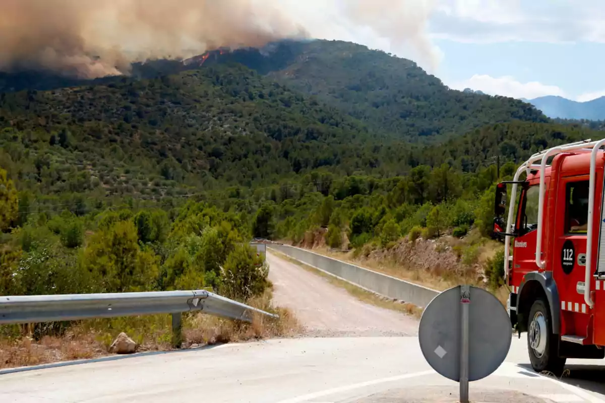 Camión de bomberos en una carretera rural mientras un incendio forestal avanza en una montaña cubierta de vegetación y humo denso en el cielo