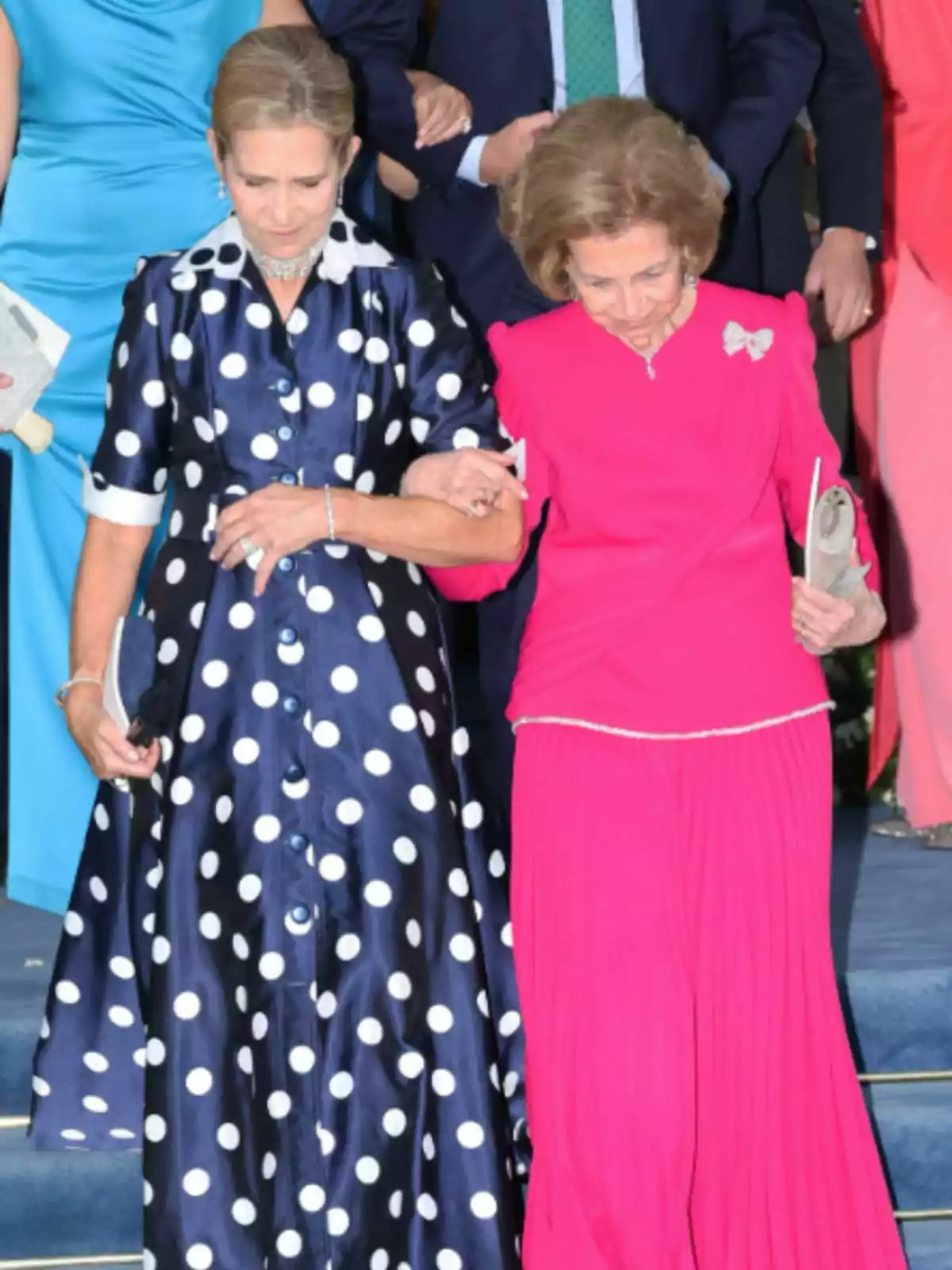 La infanta Elena y la reina Sofía elegantemente vestidas bajan unas escaleras tomadas del brazo una lleva un vestido azul marino con lunares blancos y la otra un conjunto fucsia La infanta Elena y la reina Sofía elegantemente vestidas bajan unas escaleras tomadas del brazo una lleva un vestido azul marino con lunares blancos y la otra un conjunto fucsia