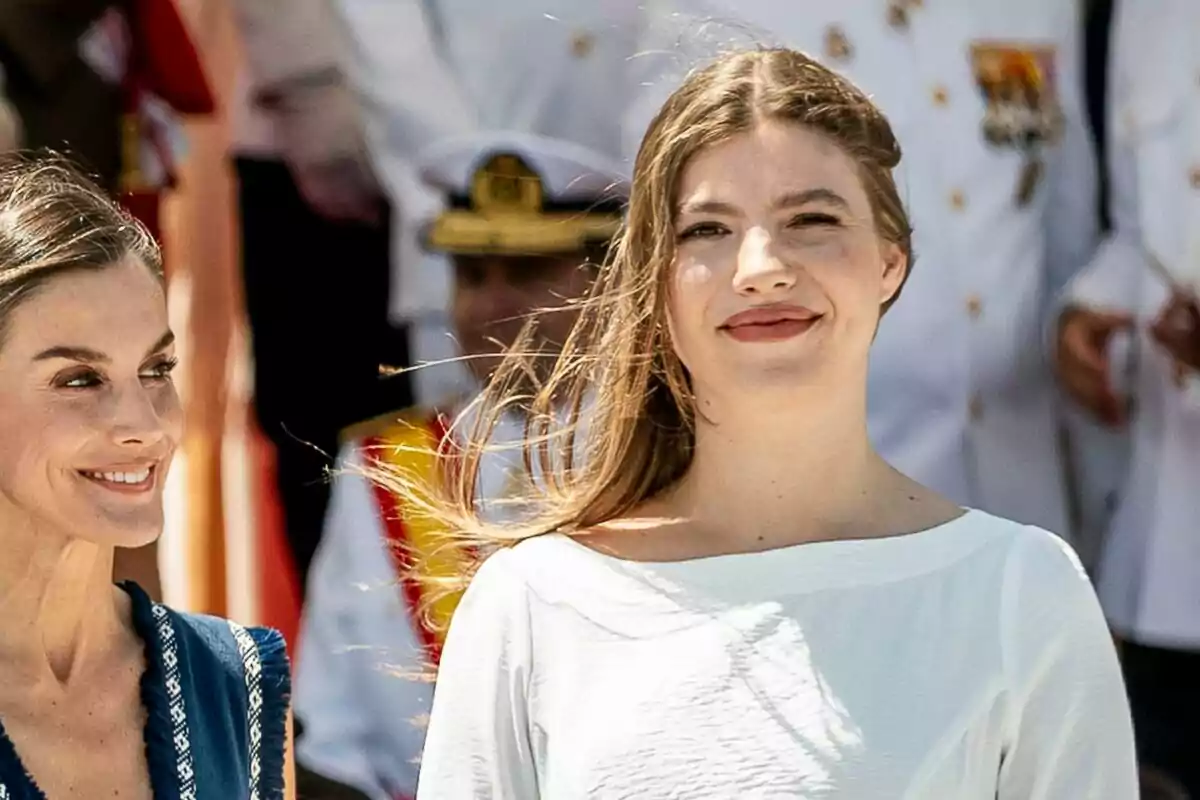La reina Letizia y la infanta Sofía sonríen al aire libre durante un evento formal con personas uniformadas al fondo La reina Letizia y la infanta Sofía sonríen al aire libre durante un evento formal con personas uniformadas al fondo