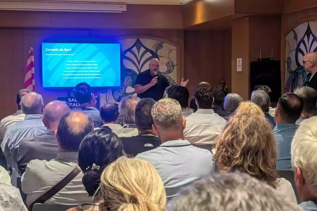 Un home dret amb micròfon parla davant d’un grup de persones assegudes en una sala mentre una pantalla blava mostra informació al fons. Un home dret amb micròfon parla davant d’un grup de persones assegudes en una sala mentre una pantalla blava mostra informació al fons.