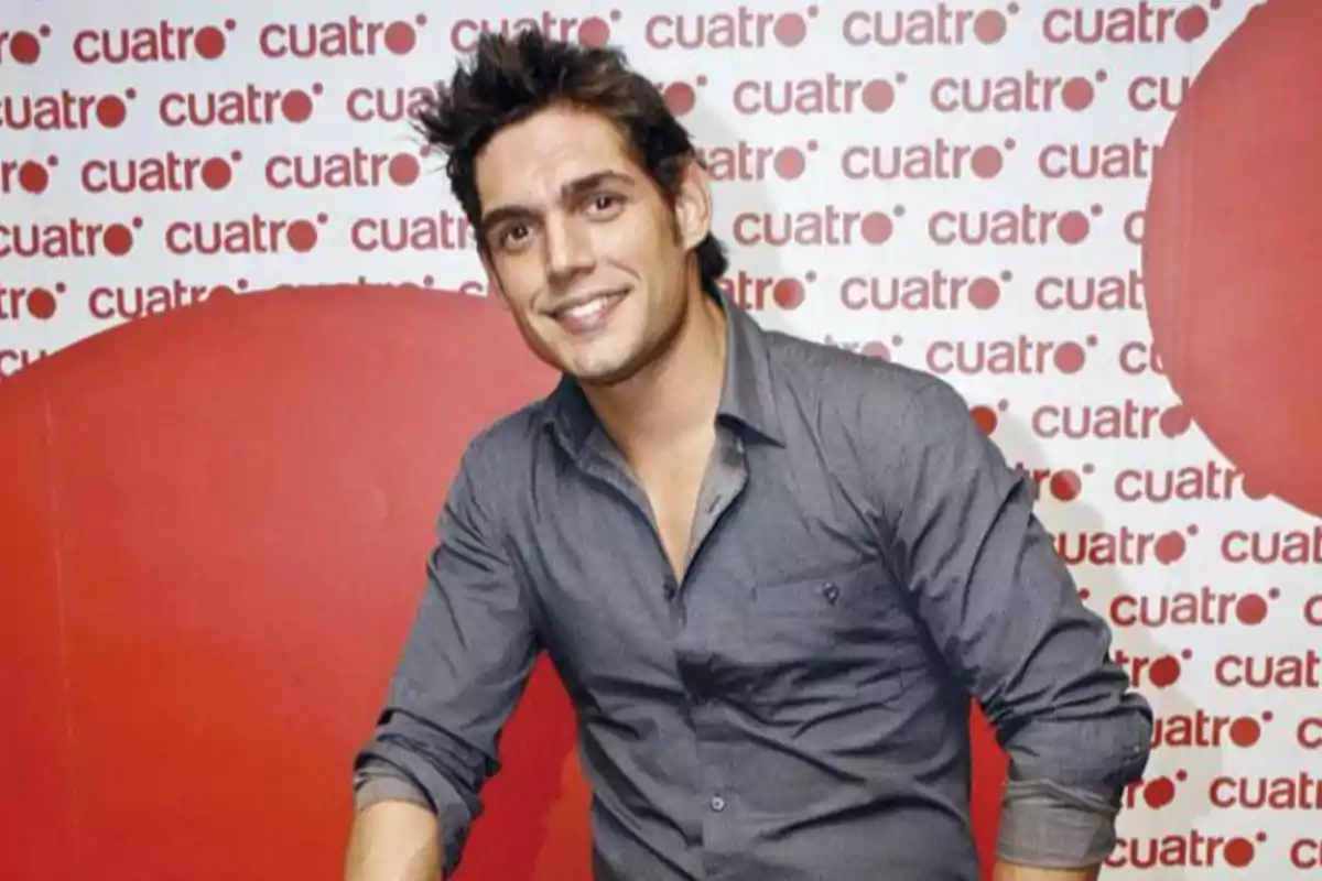 Home jove amb camisa grisa posant i somrient davant d’un fons amb el logotip de Home jove amb camisa grisa posant i somrient davant d’un fons amb el logotip de