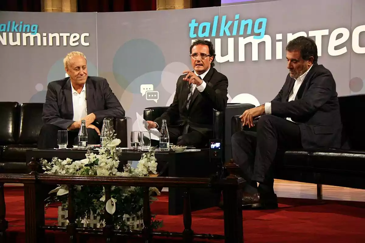Tres hombres con traje están sentados en sillones negros sobre un escenario durante un panel de discusión, con botellas de agua y flores blancas en una mesa frente a ellos y un fondo que dice talking numintec. Tres hombres con traje están sentados en sillones negros sobre un escenario durante un panel de discusión, con botellas de agua y flores blancas en una mesa frente a ellos y un fondo que dice talking numintec.
