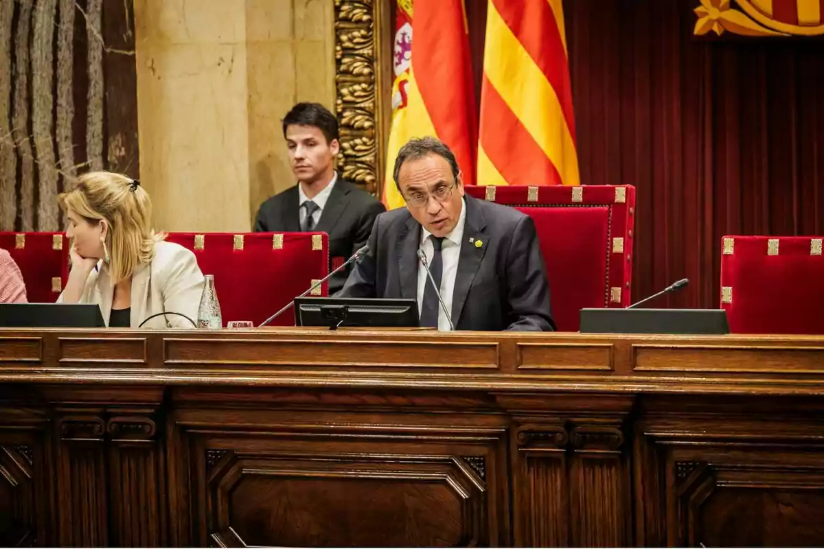 Un home amb ulleres i vestit fosc parla en un micròfon assegut en una taula de fusta en una sala parlamentària amb banderes de fons i altres persones al seu voltant. Un home amb ulleres i vestit fosc parla en un micròfon assegut en una taula de fusta en una sala parlamentària amb banderes de fons i altres persones al seu voltant.
