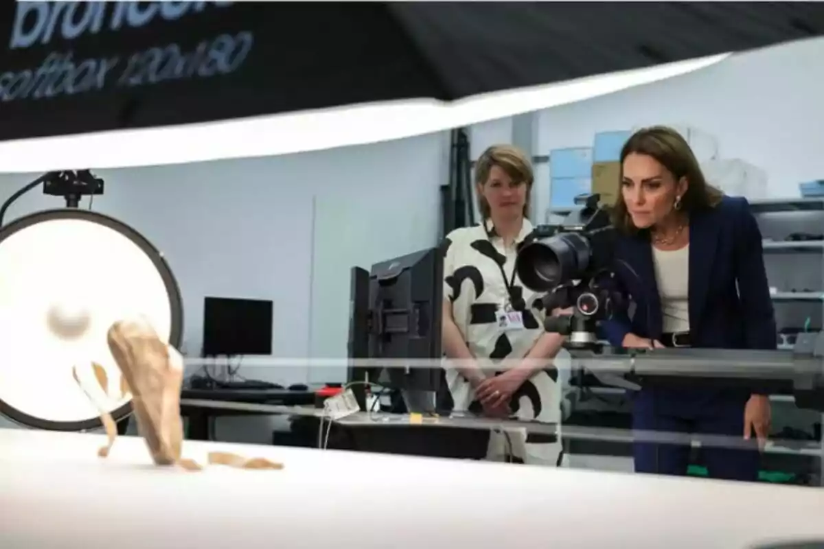 Dues dones observen de prop una càmera professional en un estudi fotogràfic mentre un objecte petit està il·luminat sobre una taula blanca. Dues dones observen de prop una càmera professional en un estudi fotogràfic mentre un objecte petit està il·luminat sobre una taula blanca.