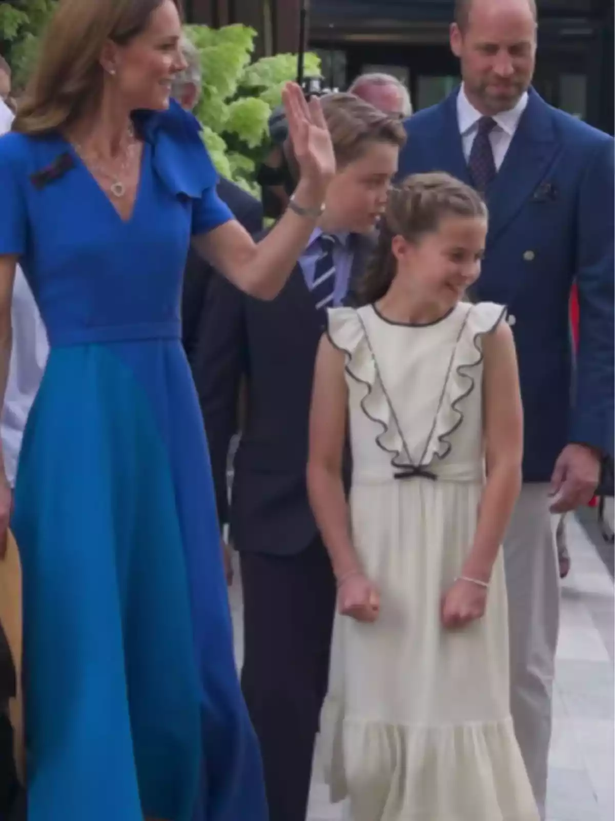 A woman in a blue dress waves while accompanied by a man in a blue jacket, a boy in a suit, and a girl in a white dress; they are all smiling and seem to be at an outdoor event. A woman in a blue dress waves while accompanied by a man in a blue jacket, a boy in a suit, and a girl in a white dress; they are all smiling and seem to be at an outdoor event.