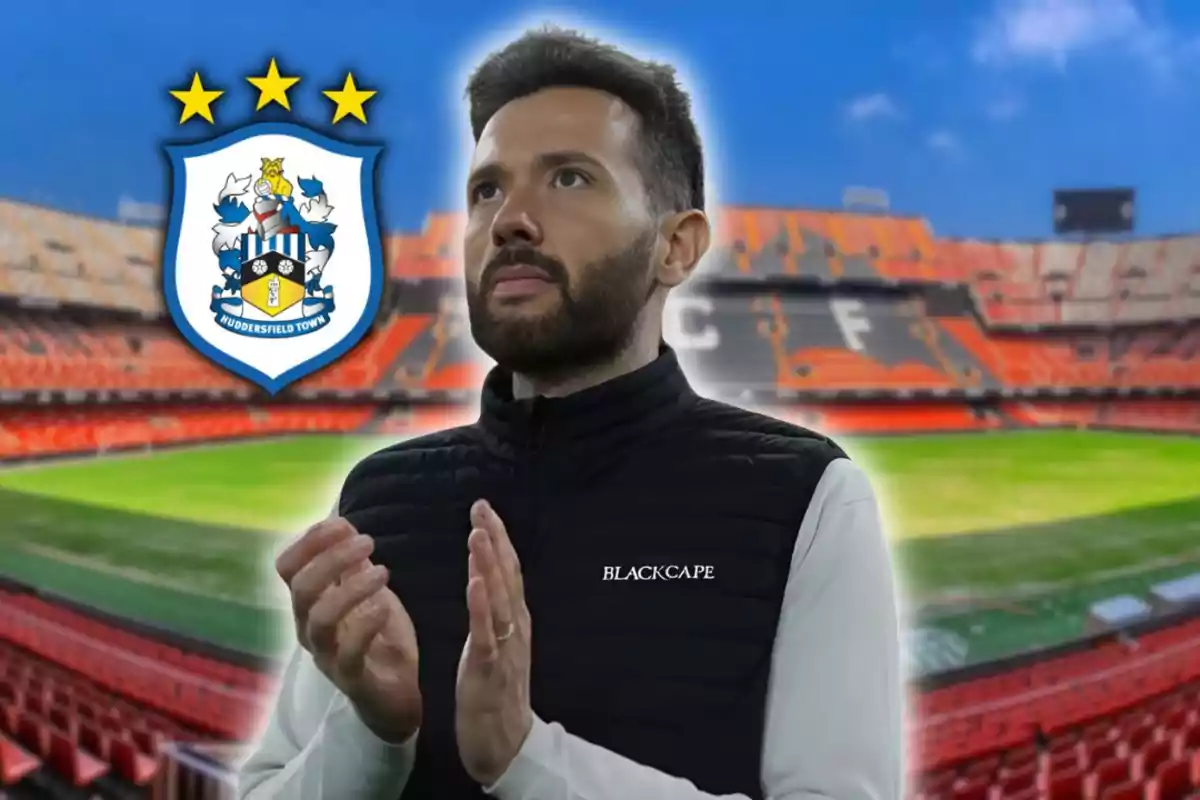 Hombre con chaleco negro aplaudiendo en un estadio de fútbol con el escudo del Huddersfield Town a su lado Hombre con chaleco negro aplaudiendo en un estadio de fútbol con el escudo del Huddersfield Town a su lado