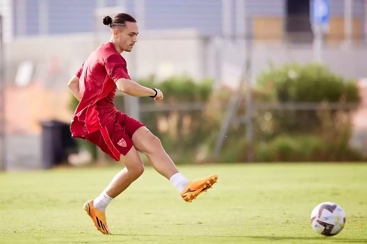 Futbolista con uniforme rojo y zapatos naranjas pateando un balón en un campo de entrenamiento Futbolista con uniforme rojo y zapatos naranjas pateando un balón en un campo de entrenamiento