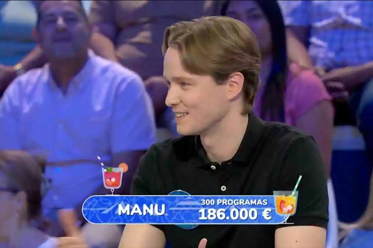 Un joven con camisa negra sonríe mientras participa en un concurso televisivo, con un rótulo que muestra su nombre, la cantidad de programas y el dinero acumulado, rodeado de público en el fondo.