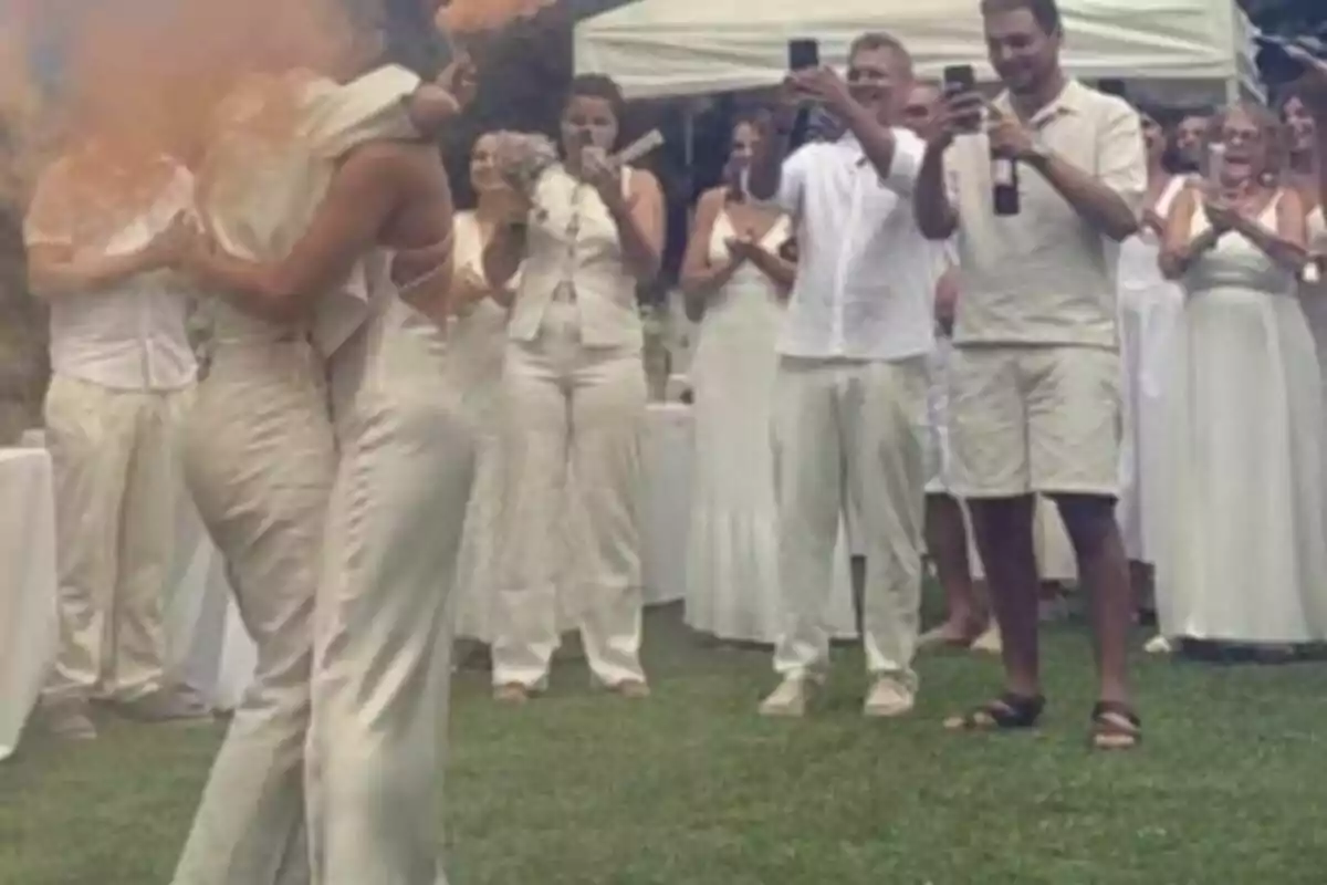 Dues persones vestides de blanc s’abracen al centre d’una celebració a l’aire lliure mentre els convidats, també vestits de blanc, aplaudeixen i fan fotos. Dues persones vestides de blanc s’abracen al centre d’una celebració a l’aire lliure mentre els convidats, també vestits de blanc, aplaudeixen i fan fotos.