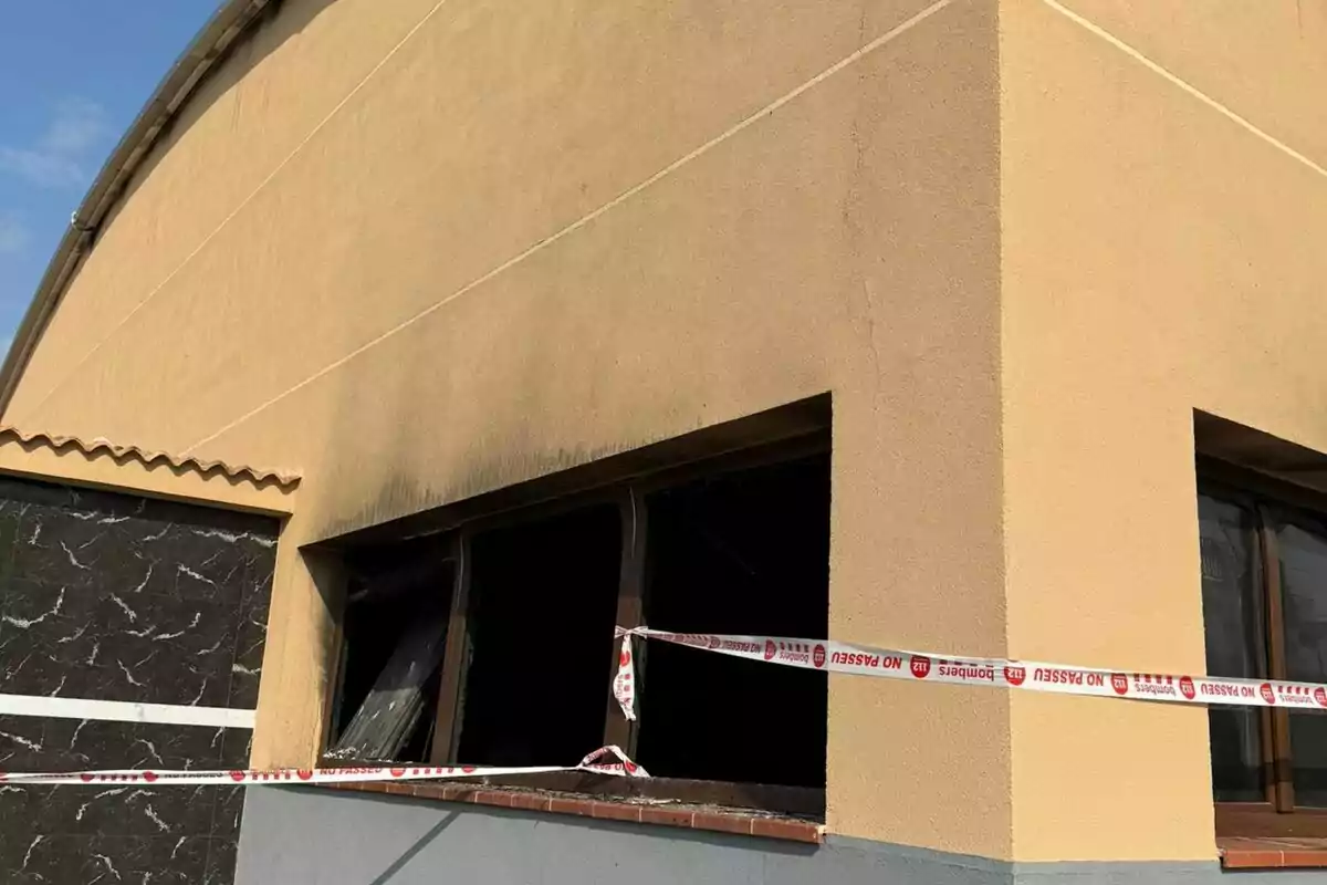 Edifici de parets grogues amb una finestra trencada i cinta de precaució de bombers envoltant la zona afectada pel fum.