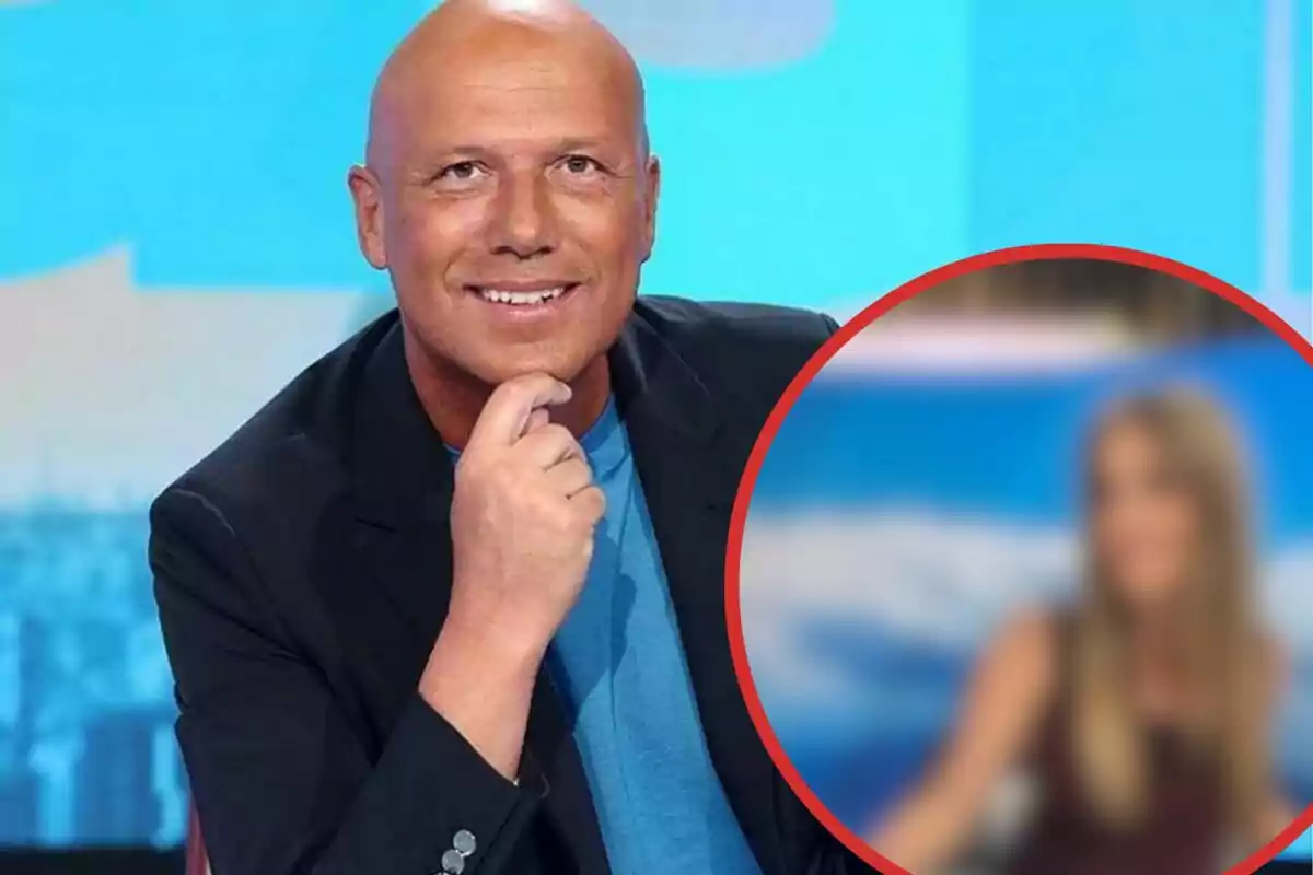 Hombre calvo sonriente con chaqueta oscura y camiseta azul sentado en un set de televisión con una figura femenina desenfocada en un círculo rojo a la derecha
