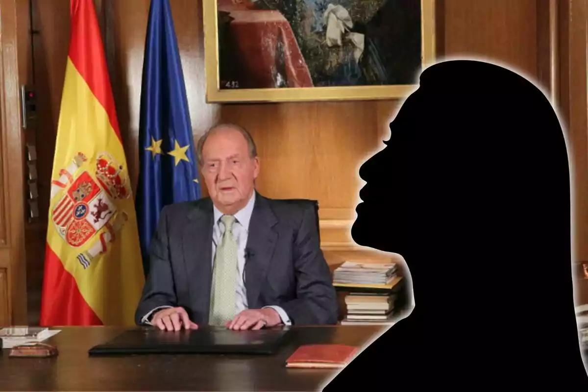 Home gran assegut en un escriptori amb banderes d’Espanya i de la Unió Europea al darrere i la silueta negra d’una persona en primer pla a la dreta