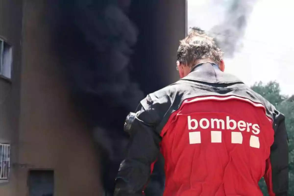 Bombero de espaldas con chaqueta roja frente a un edificio del que sale humo negro