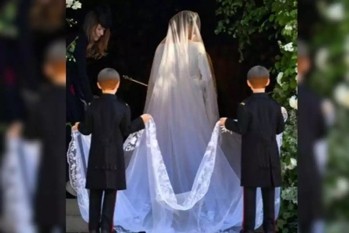 Novia vestida de blanc amb vel llarg sostingut per dos nens vestits amb vestit fosc a l’entrada d’un lloc decorat amb flors. Novia vestida de blanc amb vel llarg sostingut per dos nens vestits amb vestit fosc a l’entrada d’un lloc decorat amb flors.