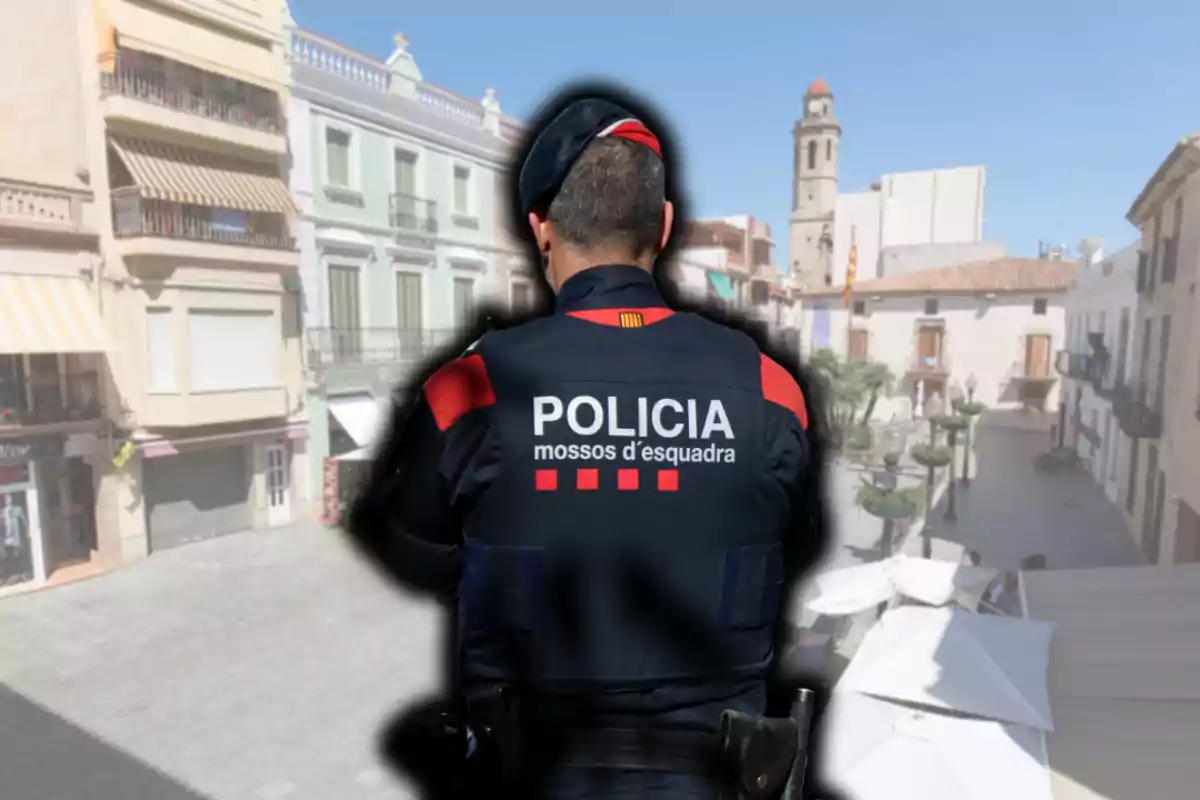 Un agent dels Mossos d'Esquadra d'esquena en una plaça d'un poble amb edificis i una torre al fons