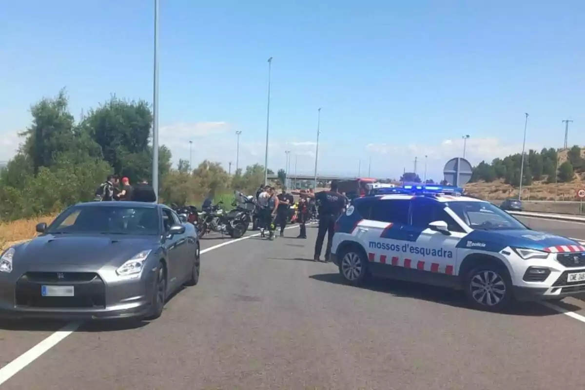 Control policial en una carretera amb diversos agents, un cotxe patrulla dels Mossos d'Esquadra, un automòbil esportiu i diverses motocicletes aturades al fons.
