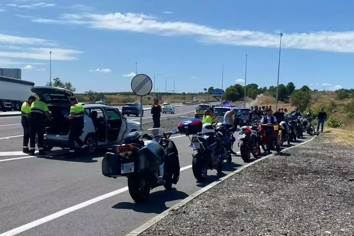 Varios policías revisan un coche detenido en una autopista mientras un grupo de motociclistas y sus motos esperan al borde de la carretera bajo un cielo despejado. Varios policías revisan un coche detenido en una autopista mientras un grupo de motociclistas y sus motos esperan al borde de la carretera bajo un cielo despejado.