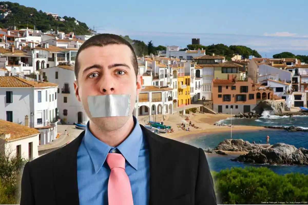 Home amb vestit i corbata rosa té la boca tapada amb cinta adhesiva platejada i es troba davant d’un poble costaner amb cases blanques i una platja