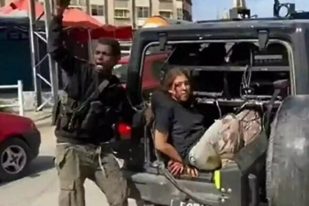 Un hombre armado sostiene la mano en alto junto a una persona herida sentada en la parte trasera de un vehículo militar en una calle urbana. Un hombre armado sostiene la mano en alto junto a una persona herida sentada en la parte trasera de un vehículo militar en una calle urbana.