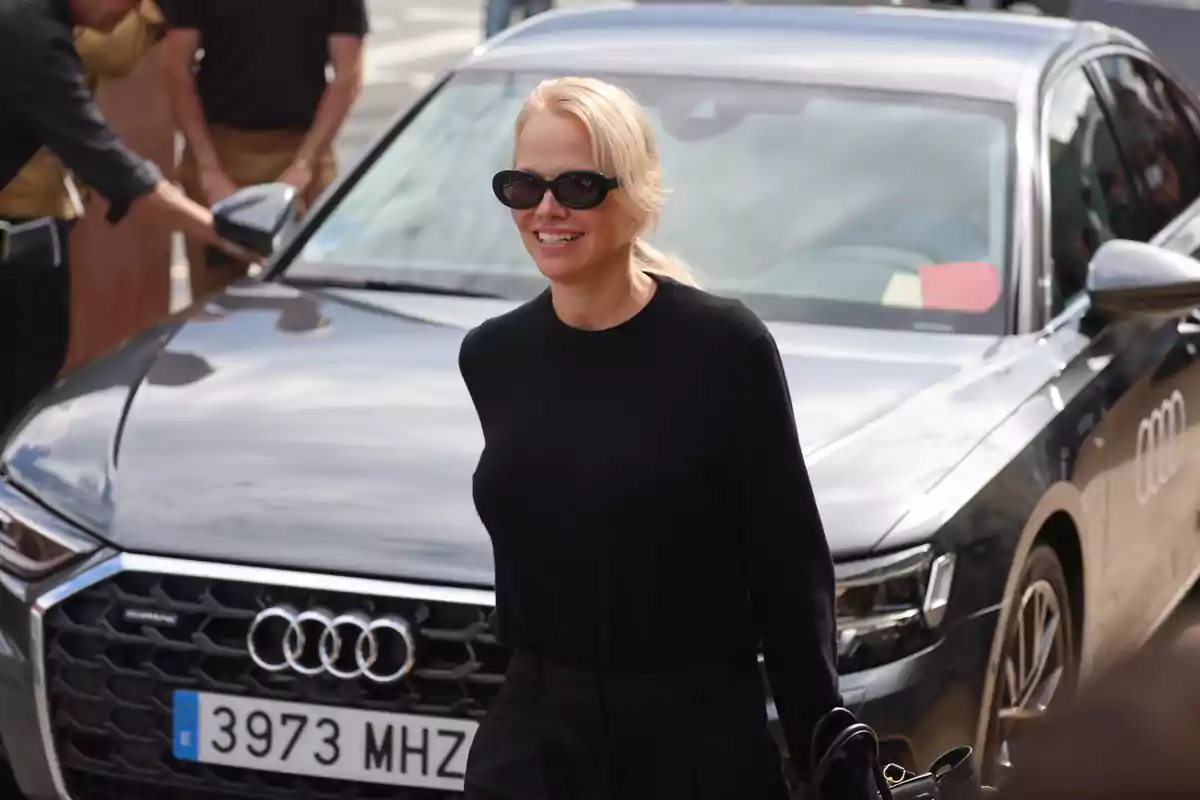 Pamela Anderson amb ulleres de sol i jersei negre somriu mentre camina davant d’un cotxe Audi platejat Pamela Anderson amb ulleres de sol i jersei negre somriu mentre camina davant d’un cotxe Audi platejat