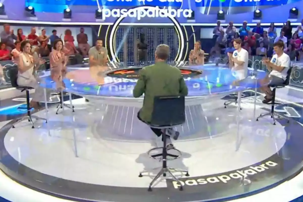 Persones assegudes al voltant d’una taula circular en un plató de televisió amb públic al fons i el logotip de Pasapalabra a terra Persones assegudes al voltant d’una taula circular en un plató de televisió amb públic al fons i el logotip de Pasapalabra a terra