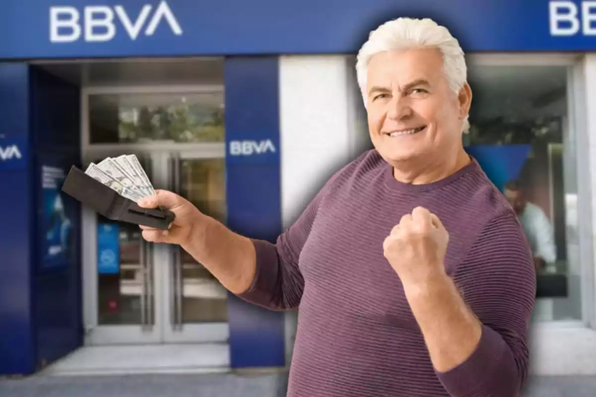 Hombre mayor sonriente sostiene una billetera con billetes frente a una sucursal de BBVA y muestra un gesto de triunfo