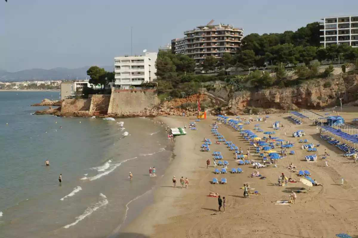 Platja amb para-sols i gandules blaves, persones gaudint del mar i edificis al fons al costat d’un penya-segat Platja amb para-sols i gandules blaves, persones gaudint del mar i edificis al fons al costat d’un penya-segat