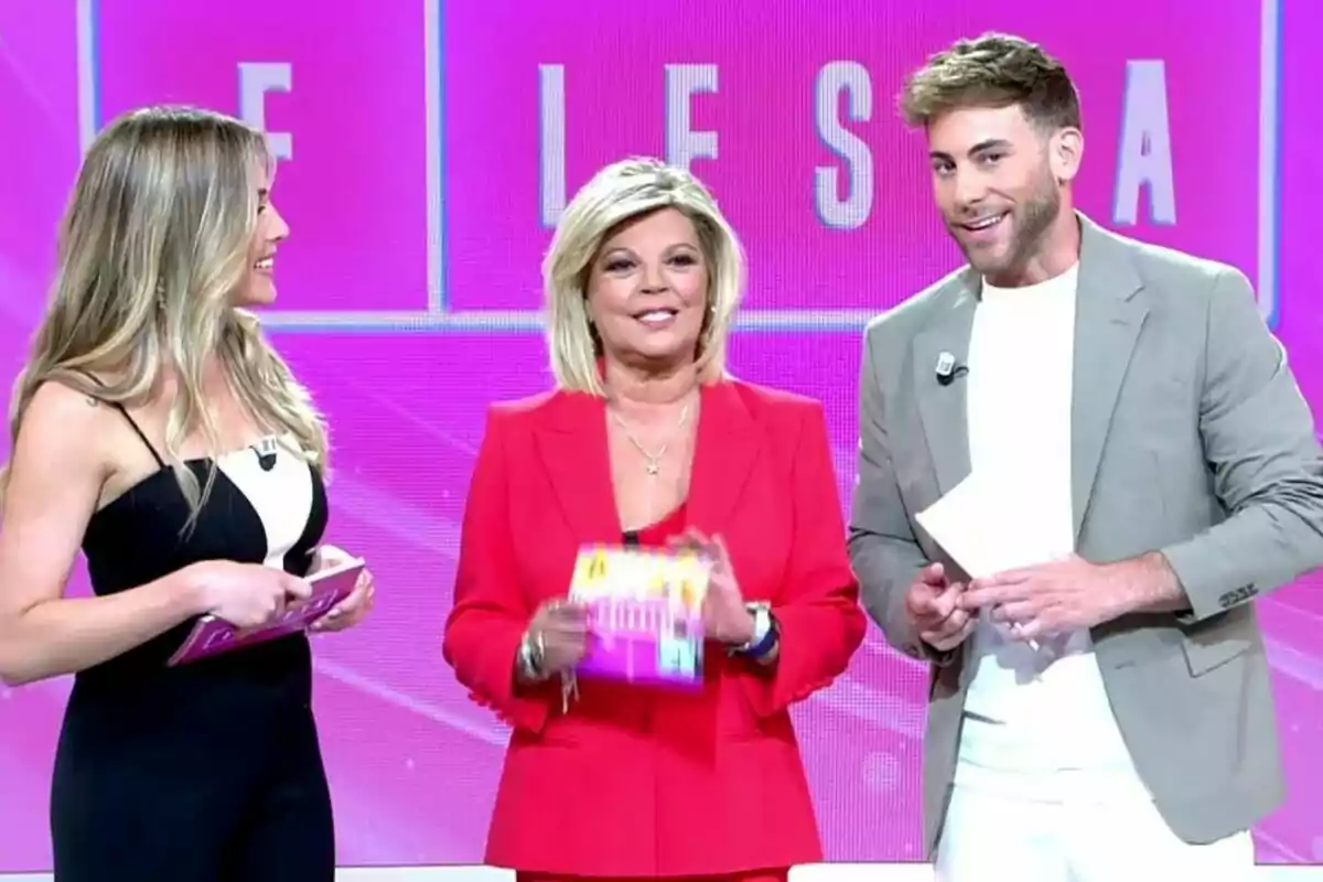 Tres presentadors de televisió, Álex Blanquer, Terelu Campos i César Muñoz, somriuen en un plató de Fiesta amb un fons rosa mentre sostenen targetes a les mans. Tres presentadors de televisió, Álex Blanquer, Terelu Campos i César Muñoz, somriuen en un plató de Fiesta amb un fons rosa mentre sostenen targetes a les mans.