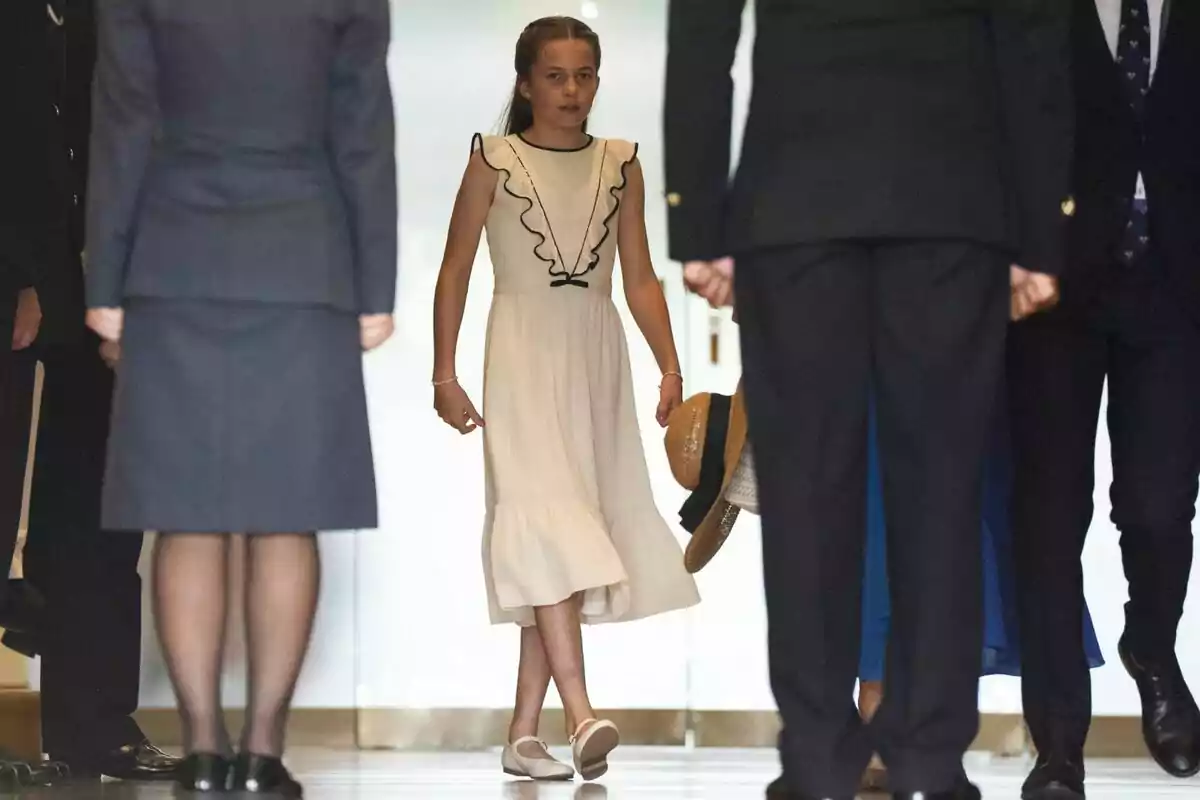 Princess Charlotte in a light dress and white shoes walking down a hallway surrounded by adults dressed formally. Princess Charlotte in a light dress and white shoes walking down a hallway surrounded by adults dressed formally.